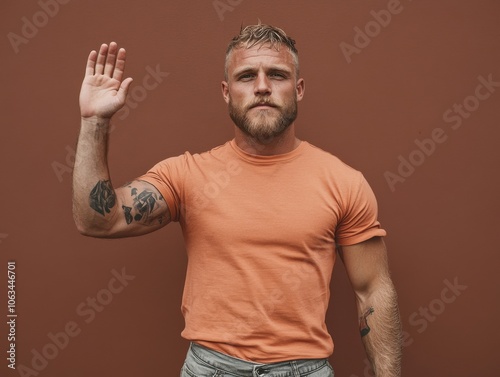 A man with a beard and tattoos raises his hand against a solid brown background, wearing a peach-colored shirt, exuding a confident and assertive vibe.