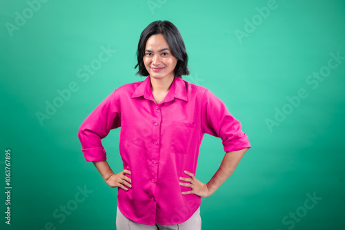 An Asian woman in a bright pink button-up shirt stands confidently with her hands on her hips against a solid green background, projecting a friendly and assertive expression