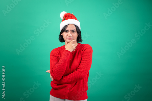 An Asian woman in a red sweater and Santa hat poses with a thoughtful expression, resting her chin on her hand, against a green background, creating a humorous and festive holiday mood