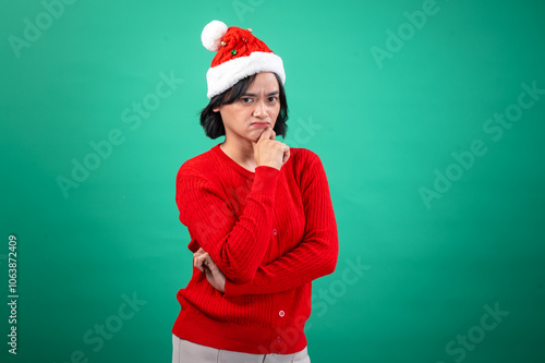 An Asian woman in a red sweater and Santa hat poses with a thoughtful expression, resting her chin on her hand, against a green background, creating a humorous and festive holiday mood