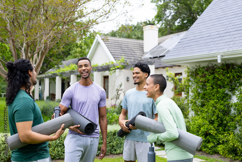 Young multiracial friends laughing and holding yoga mats outdoors in garden setting