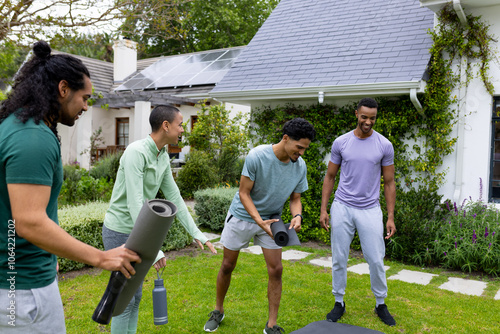 multiracial friends preparing for outdoor yoga session, smiling and holding mats in garden