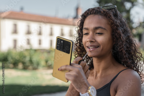 Beautiful young woman applying lip gloss while looking at smartphone screen outdoors