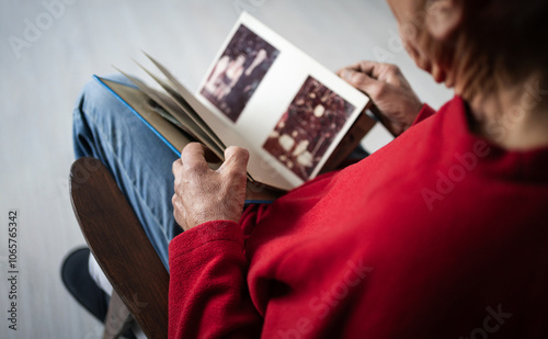 Lonely old man in red sweater sitting on chair holding photo album looking at photos remembering past and deceased wife