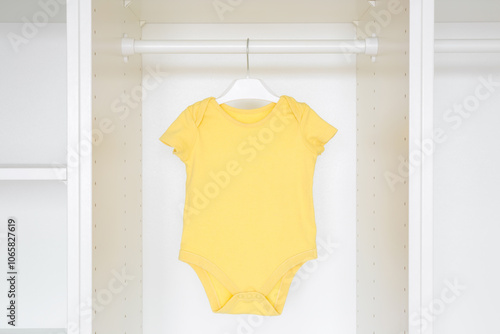 Yellow baby bodysuit hanging on hanger inside white wooden wardrobe at home. Closeup. Front view. Clothes preparing for newborn.