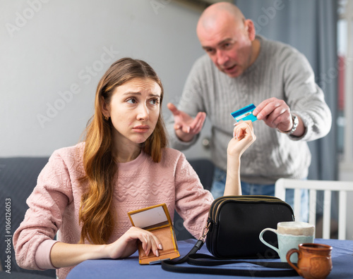 Disgruntled man picks up a credit card from a frustrated young woman sitting at a table in a room, angrily reproaching her