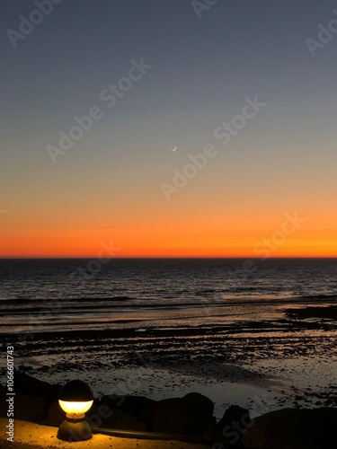 Anochecer costa andaluza, cielo arcoíris, cuarto de luna asomando tímida, horizonte naranja últimos rayos de sol, aguas mansas marea baja, contraste farola, fondo de pantalla cuadro marco