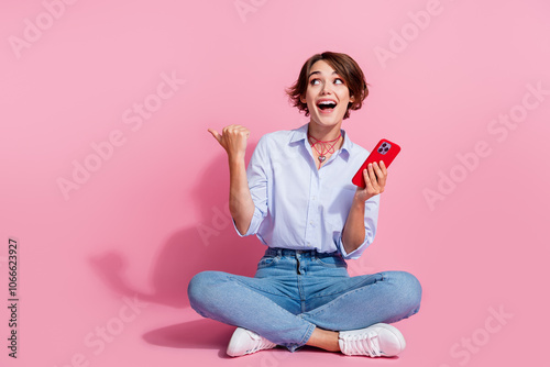 Full body portrait of charming impressed lady sit floor hold smart phone look indicate finger empty space isolated on pink color background