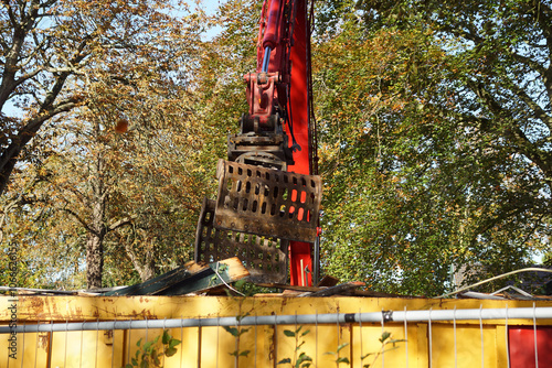 Close up excavator with grab and a rubbish skip with old wood, planks. Trees with autumn leaves on the background. Netherlands, October.