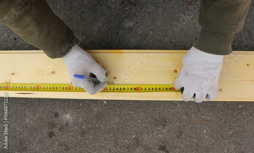 A man in work clothes and gloves uses a tape measure and pen to make markings on a light-colored wooden board lying on the asphalt