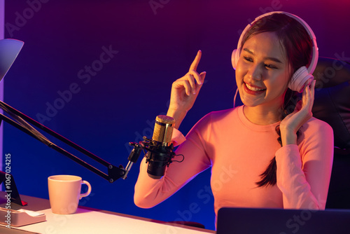 Host channel of young beautiful Asian broadcaster listening the music on social media with listeners, wearing pastel headphones, using microphone for singing at neon light indoor studio. Stratagem.