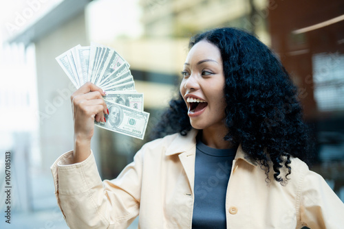 Excited young woman holding a fan of dollar bills, expressing joy and success in an outdoor urban setting.