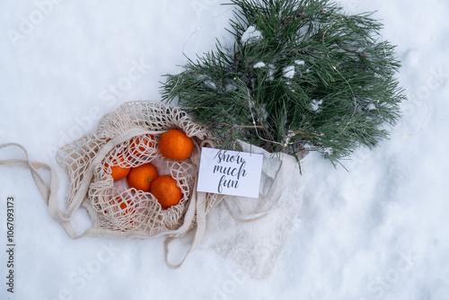 SNOW MUCH FUN text on postcard Mesh shopping bag with tangerines with fir branch and garland in snowy winter outdoor. Go green concept eco-friendly sustainable Christmas. Flat lay in snow background