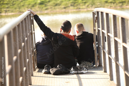 Three boys and friends are sitting on a bridge with metal handrails over the water. Friendly conversation in nature. Children's play outdoors