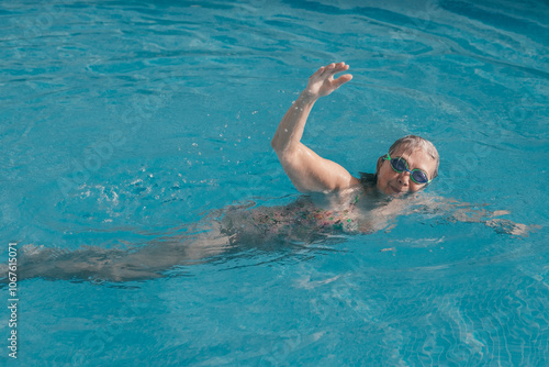 Active senior woman swimming freestyle in outdoor swimming pool under the sunlight wearing swimming goggles. Retirement and healthy lifestyle concept