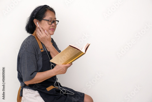 Asian housewife wearing black apron, sitting alone reading recipe book while listening to the music on her earphones