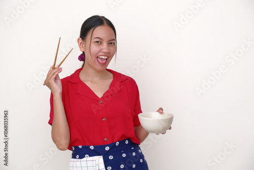 Woman in the kitchen smilingly, showing her delicious food in white bowl she is eating with chopsticks