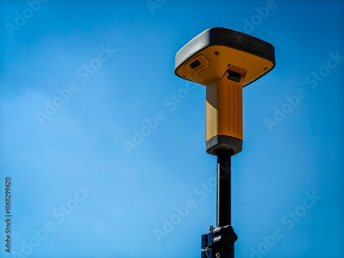 A photo of a yellow GPS receiver mounted on a tripod on a rooftop, with a lone bird flying in the clear blue sky.