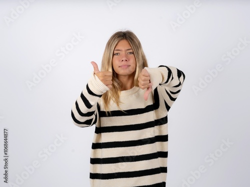 caucasian teen girl wearing striped knitted sweater over white showing thumbs up and thumbs down, difficult choose concept