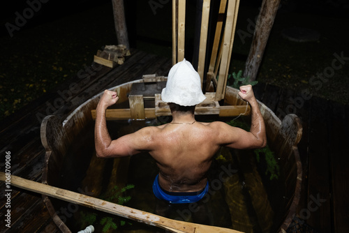 Man in wooden sauna tub flexing arms with towel hat outdoors