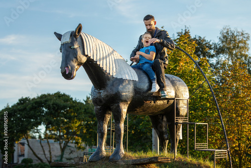 A father and son ride up a hill on a large horse sculpture. Strong emotion of a very happy child, he imagines riding a real horse.