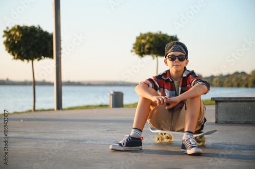 A young skater boy sits calmly on a skateboard in a bustling skate park on a sunny summer day