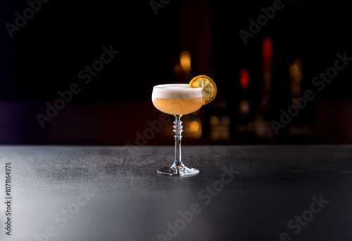 A single cocktail in a coupe glass with a lemon wedge garnish sits on a dark counter with a blurred background.