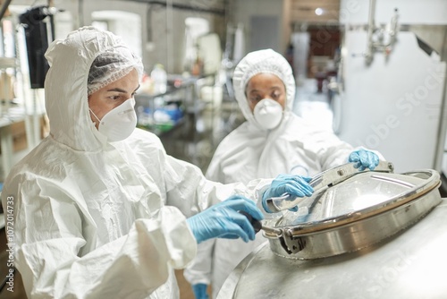 Medium shot of female process technician in protective coverall suit opening tank lid while controlling product fermentation with colleague in factory workshop, copy space