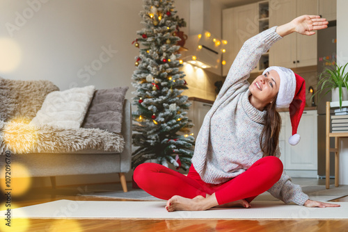 Positive woman wearing a Santa hat doing yoga on an exercise mat at home during the Christmas holidays. Holidays and healthy lifestyle.