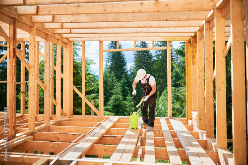 Laborer constructing wooden frame house near forest. Man treating woods, applying fire retardant using sprayer, while dressed in protective suit, helmet. Concept of modern eco-friendly construction.