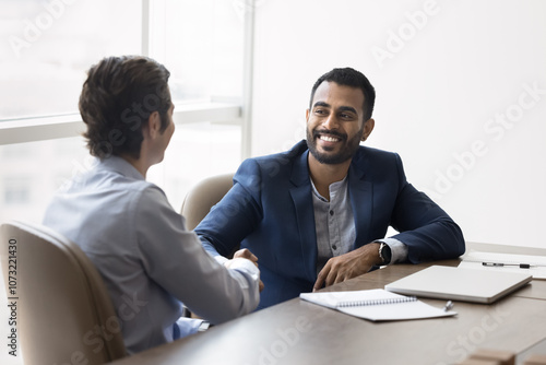 Happy young Indian business owner man shaking hands with male partner at meeting table, hiring new employee after interview, thanking financial advisor for consultation, starting partnership