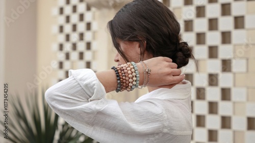 A woman in a white shirt and with bracelets on her arm stands with her back to the camera and straightens her hair.