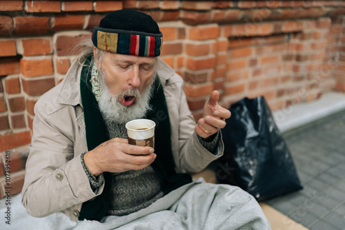 Closeup of frozen retired homeless man sitting on city street, holding hot drink to warm hands, reflecting sadness and hope. Portrait of sad aged tramp male with emotion of poverty and despair.