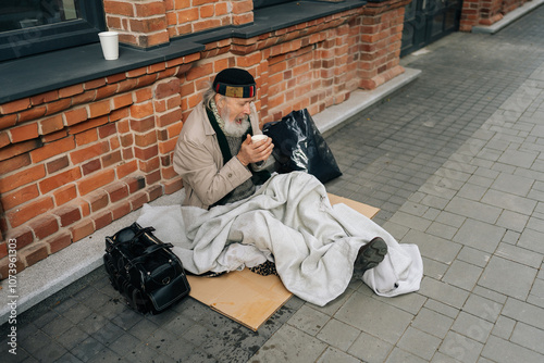 High-angle view of freezing aged homeless man sitting on city street, holding hot drink to warm hands, reflecting sadness and hope. Portrait of sad older tramp male with emotion of poverty and despair