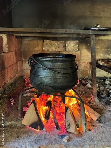 Olla de hierro fundido de tres patas sobre un fuego de leña en una parrilla. El humo y las llamas rodean la olla.