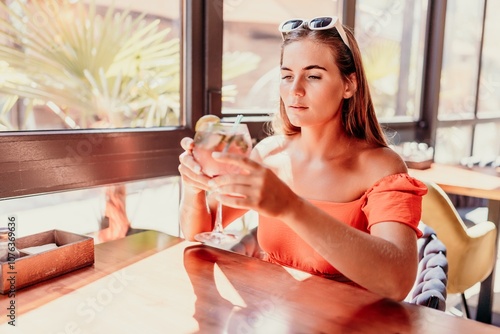 woman in a cafe seated, holding beverage. Dressed in an orange dress with her hair down. Bright interior, large windows allow natural light. Beverage consumption for refreshment.