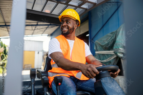 Cheerful black industry worker in working uniform driving forklift at industrial zone and looking away.