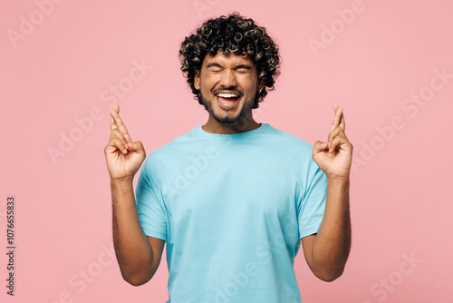 Young Indian man he wears blue t-shirt casual clothes waiting for special moment, keeping fingers crossed, making wish, eyes closed isolated on plain pastel light pink background. Lifestyle concept.