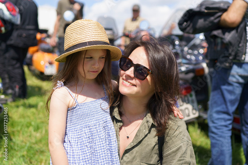 Young dark-haired mother hugs her cute daughter in a straw hat against the background of motorcycles. The concept of family values, relationships, travel and emotions