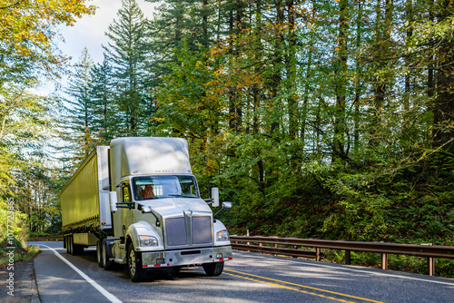 Big rig day cab white semi truck with huge roof spoiler transporting cargo in conestoga flat bed semi trailer running on the winding autumn narrow road with green and yellow trees on the sides