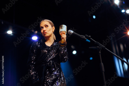 Woman Toasting with a Glass on Stage Celebrating. Cheerful party entertainer speaking at the microphone