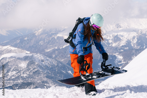 woman snowboarder wearing blue and orange ski suit and helmet fastens snowboard bindings while standing on top of slope in ski resort. Against background of grandiosely beautiful mountain range