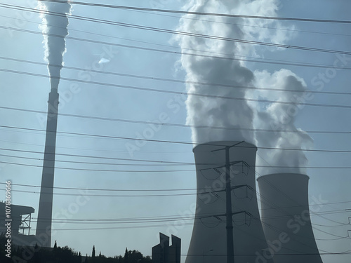 A view of large industrial towers releasing steam under clear blue skies, highlighting energy production and environmental impact. Power lines and vehicles are visible, adding to the industrial scene.