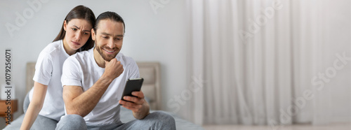 A couple sits closely on a bed, with one person smiling while viewing a smartphone. The other looks on with a concerned expression, suggesting mixed feelings in the moment.