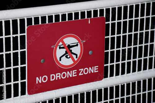 Red warning sign displaying "NO DRONE ZONE" with prohibited drone symbol mounted on white metal grid fence, close-up photograph of regulatory signage