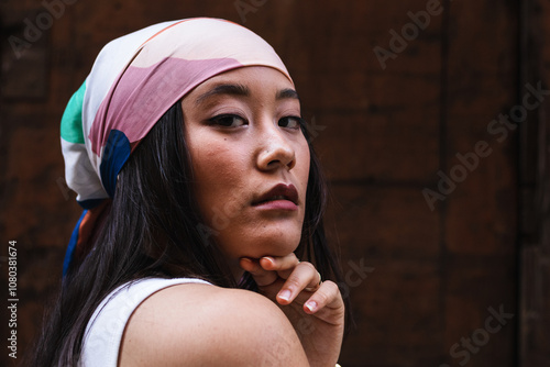 Beautiful young asian woman with headscarf posing against wooden wall, hand resting on chin in portrait