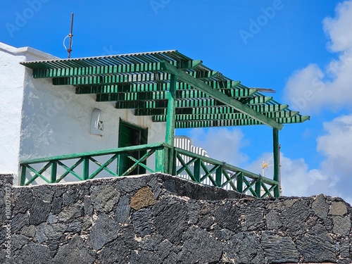 Une pergola verte ombrage une terrasse en pierres volcaniques sur fond de murs blanchis, offrant une touche de fraîcheur et de contraste sous le ciel bleu éclatant de Lanzarote.