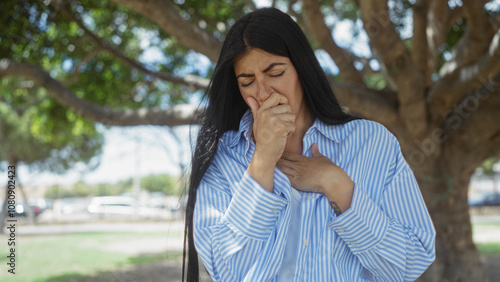 A beautiful young hispanic woman in a striped shirt stands in an outdoor park, appearing unwell as she covers her mouth and touches her chest with a concerned expression.