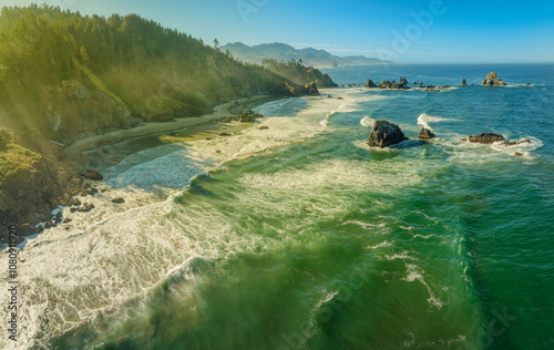 A stunning coastal landscape featuring green waters, rocky formations, and a forested backdrop under a clear blue sky. The waves crash dramatically onto the rugged shoreline.