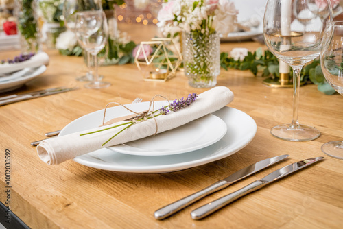 A rustic yet refined table setting featuring a white plate with a lavender sprig-tied napkin, polished silverware, and glassware. Floral arrangements and a geometric gold candle holder add elegance to